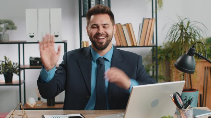 在笔记本电脑前工作的商人对着镜头友好地微笑，在办公室里挥手打招呼
