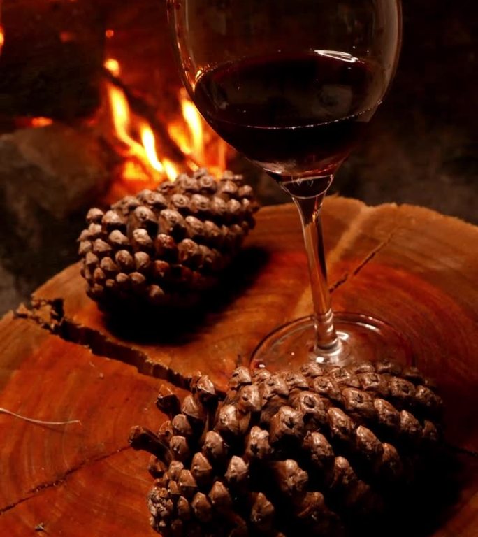 POV:在壁炉旁观察一杯酒的人。木桌上的一杯红酒。壁炉旁的一杯酒。在壁炉里燃烧的木头。喝葡萄酒。在壁