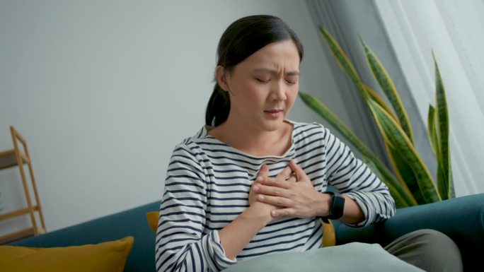 一位亚洲妇女坐在家里客厅的沙发上感到胸痛。
