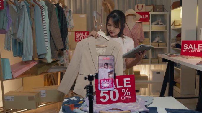 年轻的亚洲女子在晚上通过手机直播向商店里的顾客展示最新的时装系列。中小企业小企业电子商务。