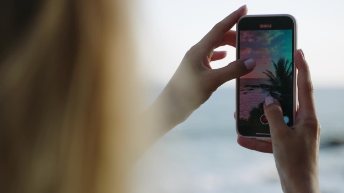 女子手持智能手机，记录热带岛屿的自然风光。手机屏幕上有蓝色的岩石海洋，蓝色的多云天空和绿色的棕榈树景