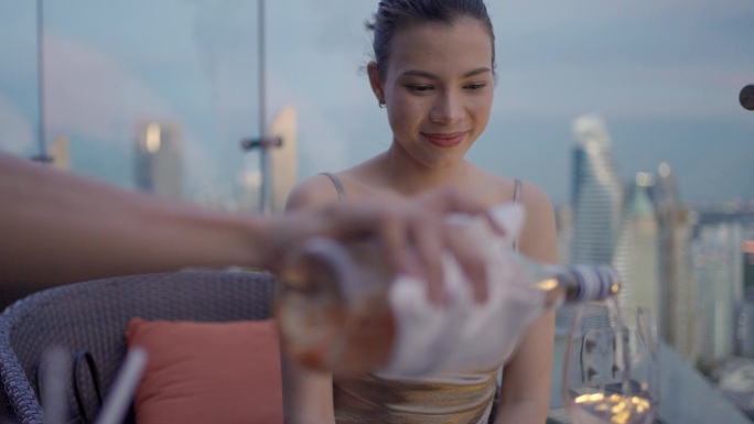 在一家城市酒店的屋顶酒吧里，男服务员拿着一瓶粉红色的葡萄酒，为一位优雅的女士斟酒。