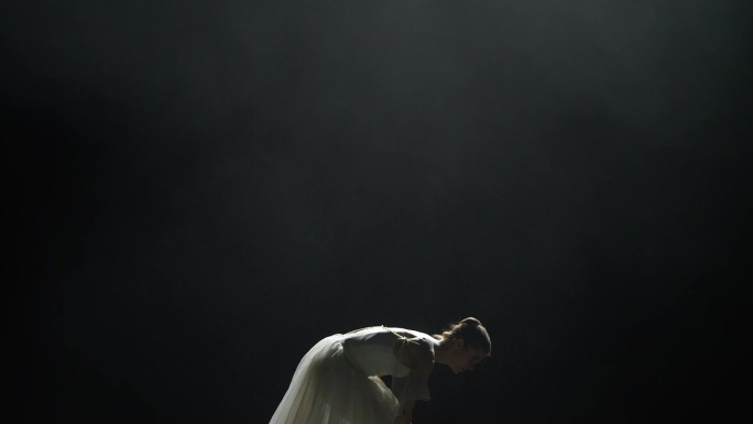 戏剧性的舞蹈，优雅的女人穿着白色的芭蕾舞裙，在黑色的背景上表演舞蹈元素，在黑暗的舞台上使用镁砂，薄雾