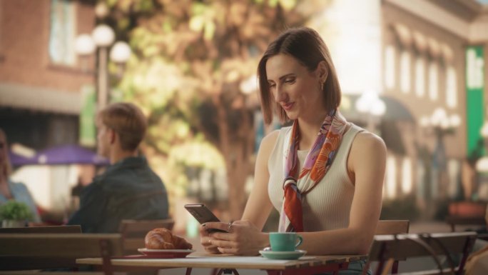 一个快乐的女性在餐厅露台上使用智能手机，享受着一杯咖啡。年轻女子在收到短信时微笑，在网上与朋友聊天，