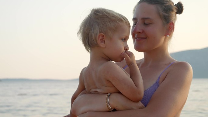 夕阳下的沙滩上，穿着比基尼的微笑女子抱着她的儿子。假期、暑假、旅游。