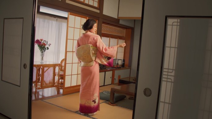 日本女性温柔地看着镜头，温柔地微笑，穿着传统的粉红色和服，上面饰有花。年轻漂亮的女人在家里的客厅里跳