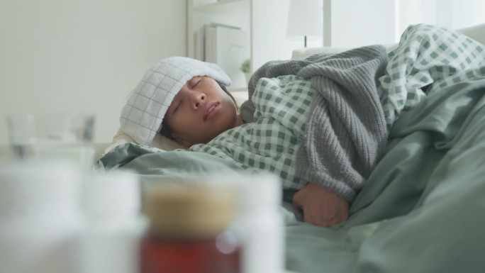 生病的年轻亚洲妇女头痛发烧咳嗽感冒打喷嚏躺在家里客厅沙发上的毯子下。医疗、居家隔离、社交距离。