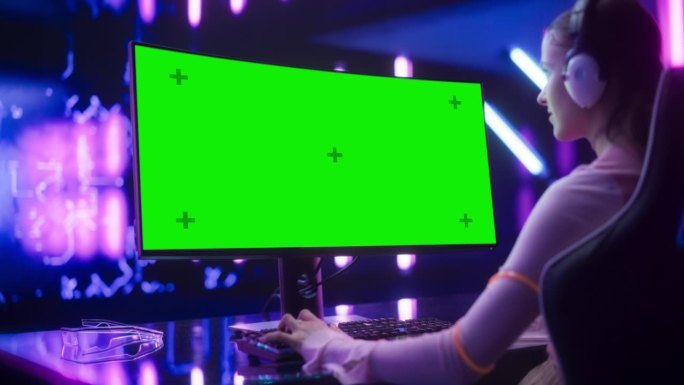 一个年轻的兴奋的玩家玩视频游戏在台式电脑上与绿屏模拟显示的镜头。年轻女性在未来的霓虹灯房间里专注于在