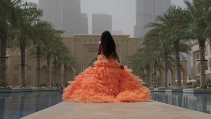一个身穿粉红色长裙、皮肤黝黑的年轻女子沿着喷泉的边缘从镜头前走了出来
