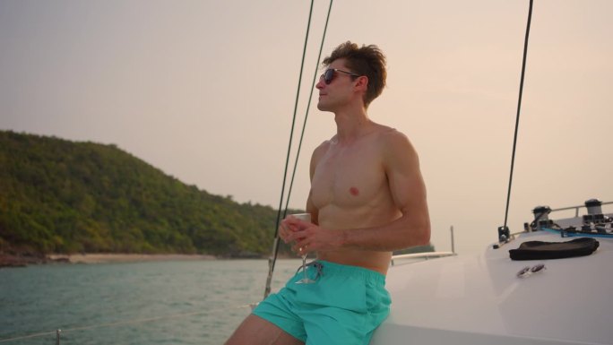 亚洲帅哥坐在游艇甲板上喝香槟。迷人的男性游客独自闲逛，庆祝假期旅行，而双体船在夏日夕阳下航行。