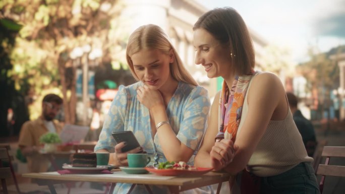 周末，两个年轻女子坐在一家餐厅的户外露台上放松。女性朋友在智能手机上聊天，浏览互联网和在线社交网络