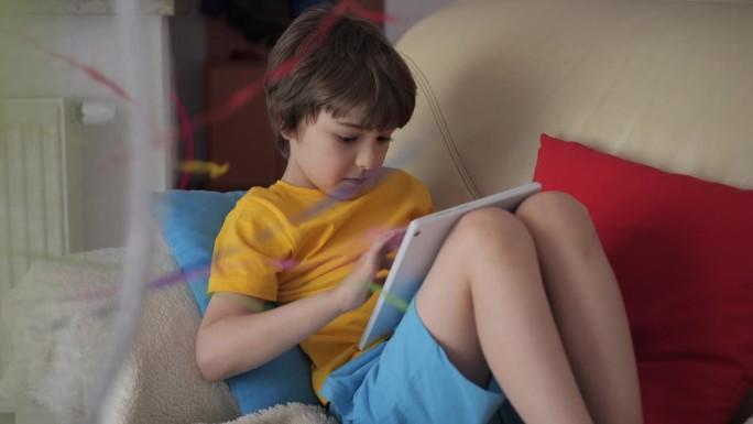 在炎热的夏天，男孩坐在沙发上，坐在风扇旁边，用平板电脑浏览社交媒体。孩子在炎热的天气里在风扇旁玩平板