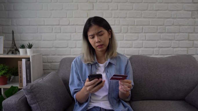 一名亚洲女子坐在沙发上，手里拿着信用卡，用手机试图通过银行应用程序付款。由于资金不足，她遇到了问题。