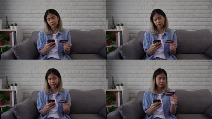 一名亚洲女子坐在沙发上，手里拿着信用卡，用手机试图通过银行应用程序付款。由于资金不足，她遇到了问题。