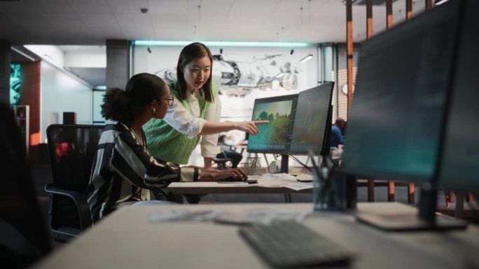 黑人女性游戏设计师使用带有3D建模软件的台式电脑与游戏开发工作室办公室的亚洲女性项目经理交谈。不同的