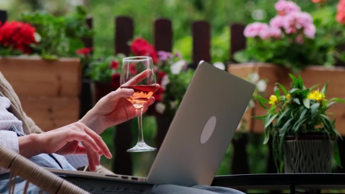一个女人手拿着一杯玫瑰酒，在舒适的家庭露台上使用笔记本电脑。女性的手指在触摸板上滑动。家庭办公，远程