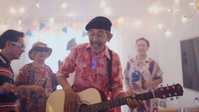 微笑的老人弹吉他庆祝他的生日，而他的朋友们在家里唱歌跳舞
