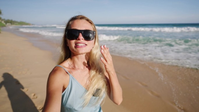 视频博主女孩用自拍相机拍摄自己，看到海洋、沙滩和棕榈树。戴着墨镜的女旅行者走在岸边，记录着在热带岛屿