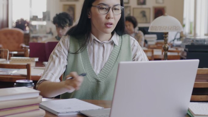 开朗的亚洲女学生在大学图书馆用笔记本电脑打字、浏览、记笔记和写笔记