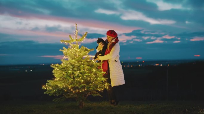 黄昏时分，一名妇女在户外欣赏圣诞树时，手里拿着一只猫
