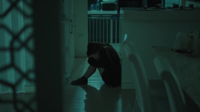 一个亚洲女人晚上独自坐在厨房的地板上，膝盖翘起，感到非常绝望，她把脸埋在膝盖上，默默地哭泣。