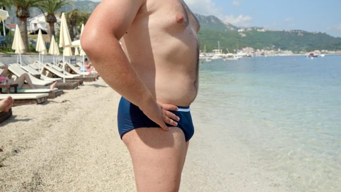 一个肥胖的年轻人在海边的特写，在海滩游泳前花了一点时间来管理他的肚子，这是他夏天前健康和体重管理的一