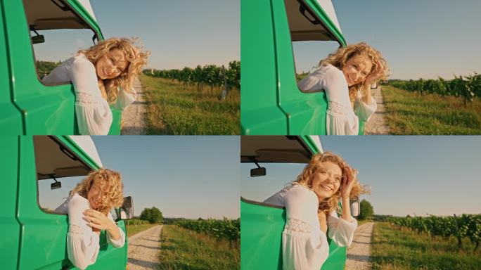快乐的金发女子探出汽车之家的车窗，拥抱着风景秀丽的乡村道路