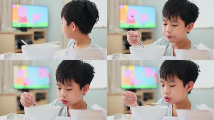 年轻的亚洲病人在流感康复期间享用营养早餐和看动画片。男孩舒服地坐在病床上，端着一盘食物，包括一碗粥和