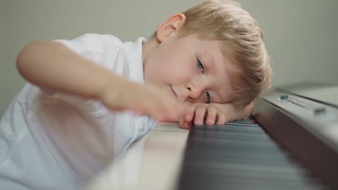 可爱的男孩把头靠在钢琴上，懒洋洋地沿着琴键移动手指