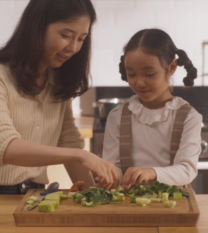 竖屏:韩国妇女和女儿在厨房一起做饭的肖像。妈妈教她的小女孩如何切蔬菜。小女孩帮妈妈准备午餐