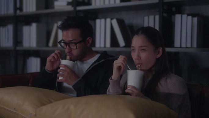 一对年轻的亚洲夫妇坐在家里的沙发上，一边吃着方便面，一边看着电视上的电影，聊得很开心。