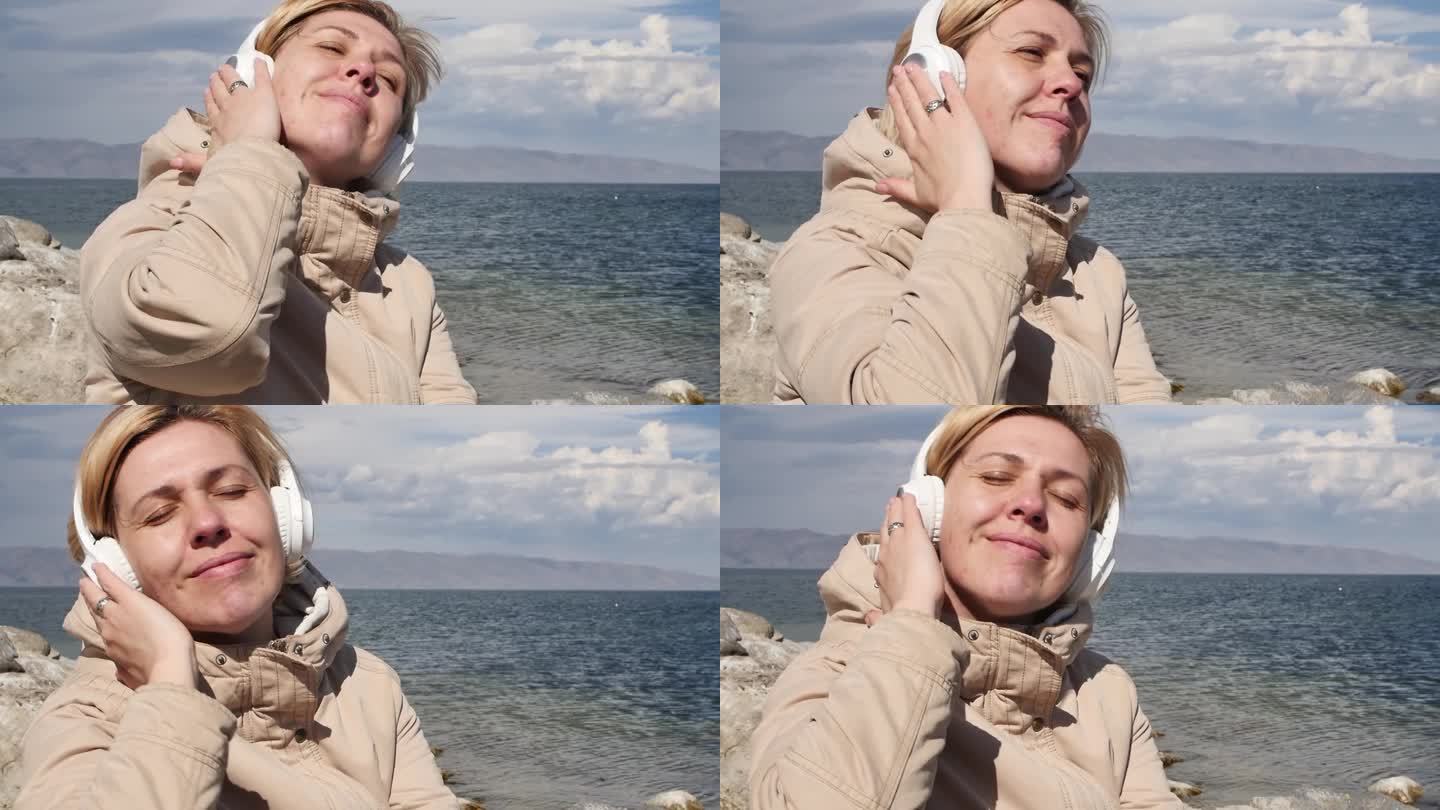 一个快乐的成年金发女子坐在海边，戴着白色的无线耳机听音乐，可以看到岸边的山峰和飞翔的海鸥享受生活