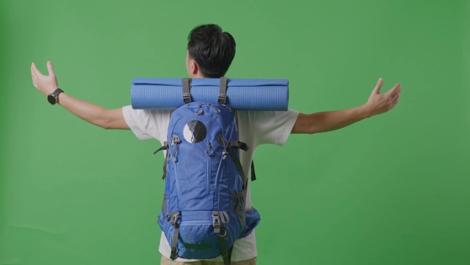 一名男性徒步旅行者背着登山背包，伸开双臂，站在摄影棚的绿幕背景上，向四周眺望