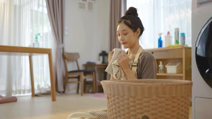 年轻的亚洲女子坐在地板上在洗衣房叠衣服，女孩从篮子里拿出衣服，叠好，闻闻洗衣机里的衣服，在一堆家务中