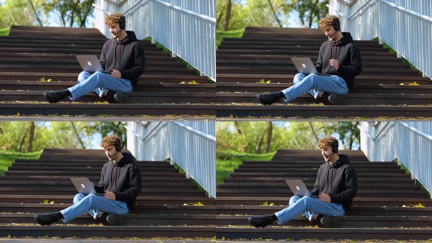 快乐的男接线员坐在公园的楼梯上，一边用笔记本电脑和耳机打视频电话。秋天的季节。教育，工作理念。慢动作