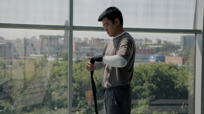 亚洲男子自由搏击运动员准备战斗的肖像，站在轻型健身房，用绑带包扎手