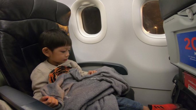 一个小男孩坐在飞机的座位上，盖着毯子，在飞机着陆时看到窗户