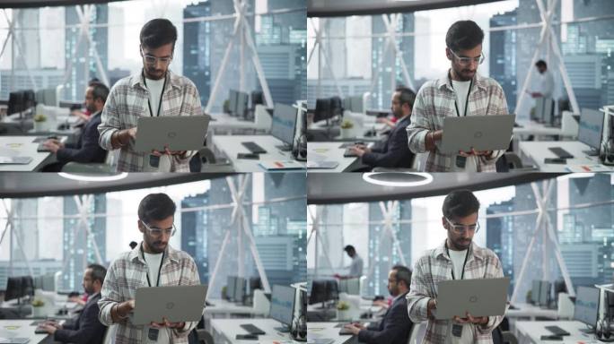 专业的印度专家站在靠近窗户的办公室里使用笔记本电脑，可以看到大城市的景色。成功的软件数据工程师优化企