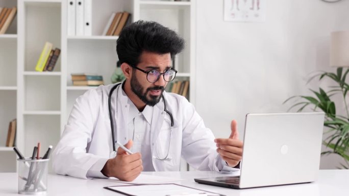 穿着白大褂的聪明友好的印度男性临床医生在医院办公室用笔记本电脑远程咨询病人。远程医疗。网上预约医生。