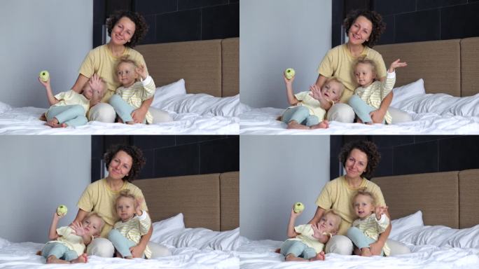 妈妈和两个女儿坐在床上微笑着看着镜头。女孩们对着镜头挥手致意或道别。视频聊天与快乐的女人与孩子在床上
