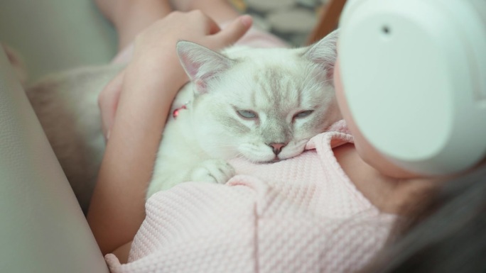 这只白色的小猫咪被她的小女孩主人抱在怀里，小女孩正躺在客厅的沙发上，听着耳机里的音乐休息。小孩和宠物