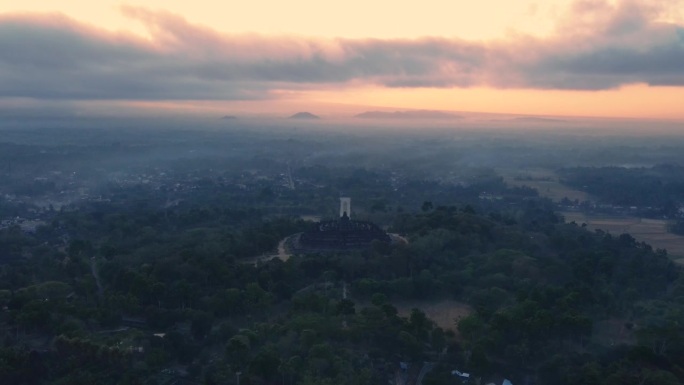 揭示无人机拍摄婆罗浮屠寺与神奇的日出光在雾蒙蒙的早晨。大地仍然一片漆黑，天空是橘黄色的。古老的佛教寺