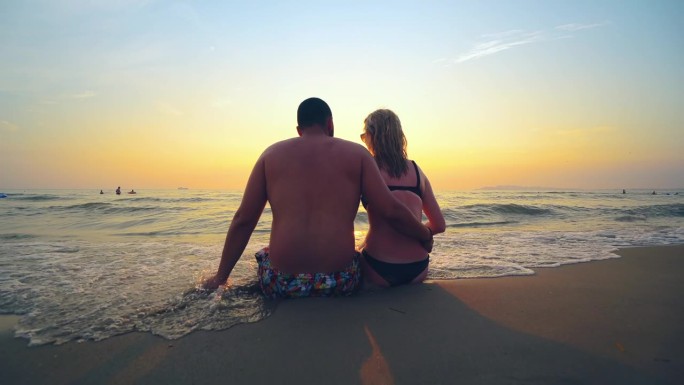 夏天旅行、度假时，一对相爱的情侣在海滩上一起看日落。剪影从后面坐着拥抱享受日落，日出的海洋上的热带目