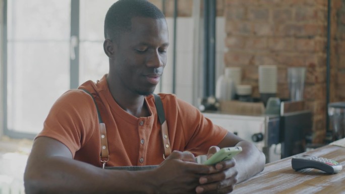 非洲裔美国男咖啡师站在咖啡馆的柜台前，微笑着用智能手机在社交媒体上聊天