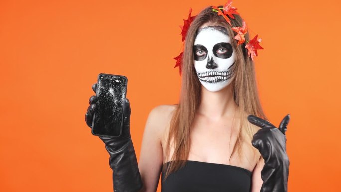 概念，万圣节的漂亮妆容。一个年轻性感女孩的骷髅妆肖像。在橙色背景下，他指着手机。
