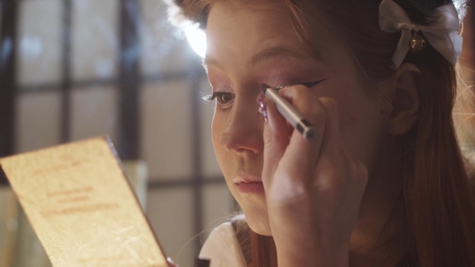 一个可爱的女孩正在化妆，画着戏剧性的双翼眼线，看着小镜子，涂着眼线液