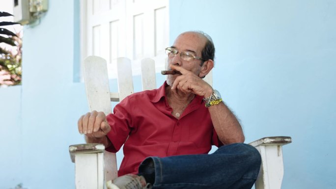 西班牙裔老人在自家门廊放松，一边抽着传统的古巴雪茄一边向邻居打招呼
