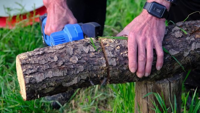 手持便携式野营电锯上的电池，用于切割柴火和木材。特写，一只手在锯木头，碎片四处飞舞