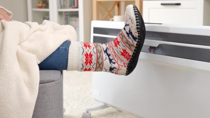 穿着针织袜的妇女在电热器前暖脚的特写。能源危机的概念，高账单，节约和节省每月的公用事业费用。
