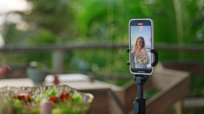 智能手机屏幕上，年轻女博主在热带岛屿的咖啡馆里用蔬菜沙拉自拍视频。三脚架上的手机拍下了女性旅行者在户